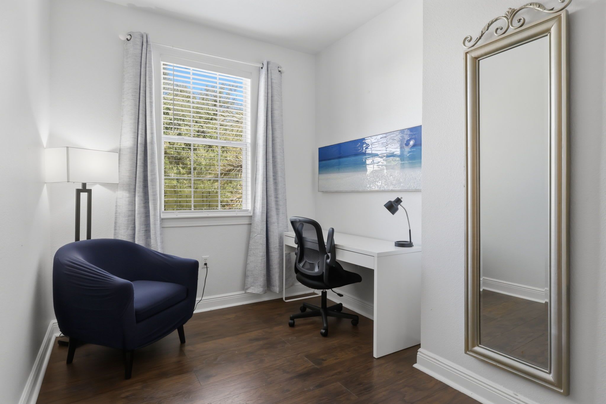 A room with a desk, chair, lamp, window with curtains, and a large mirror.
