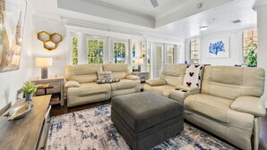 A spacious living room with a beige sofa set, a grey ottoman, and a wooden coffee table. There are multiple windows providing natural light, and a ceiling fan is visible. - New! Poolside Reunion Retreat Near Orlando Attractions (Kissimmee)