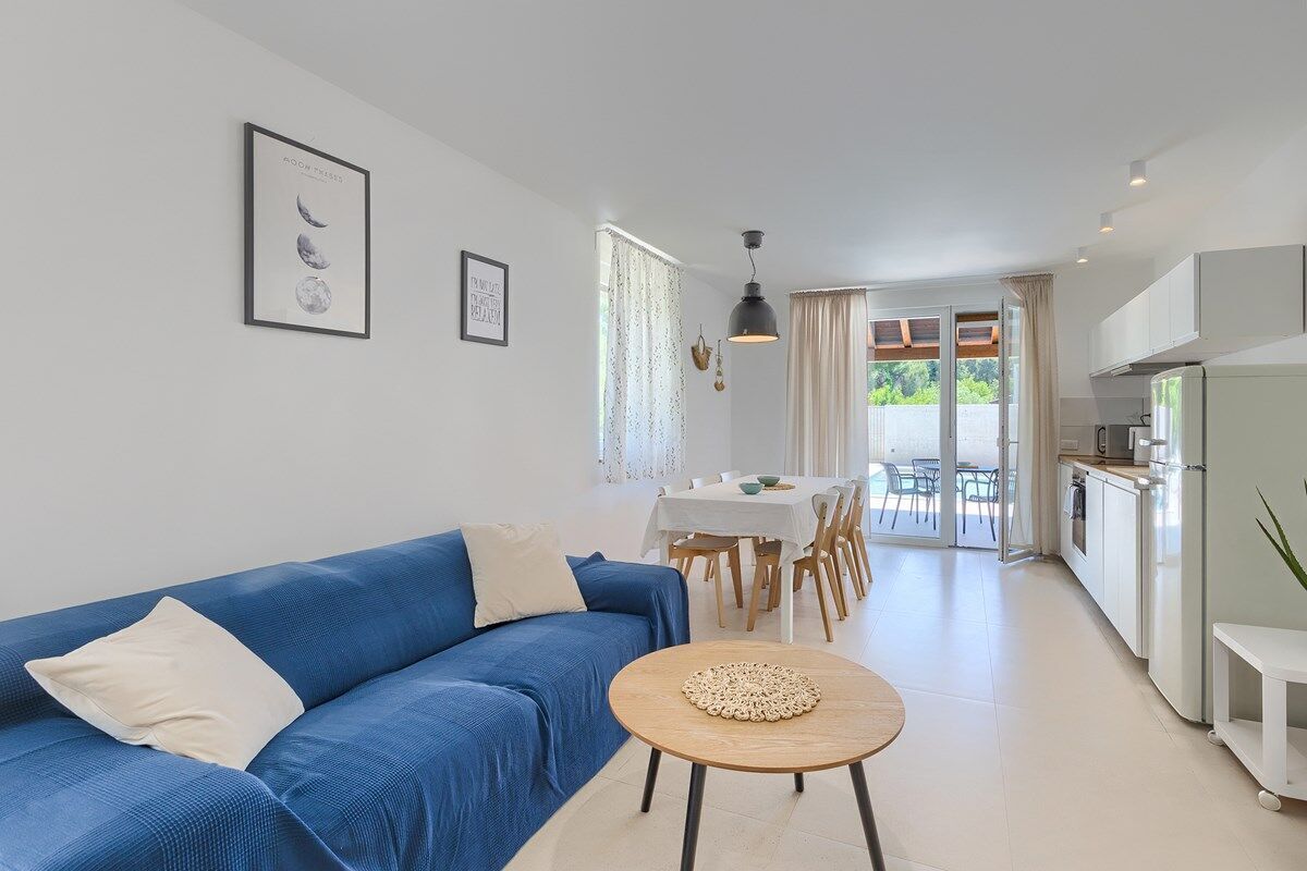A modern living room with a blue sofa, a round wooden table, and a dining area with a white tablecloth.