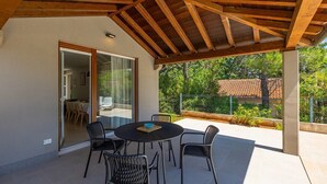 Een terras buiten met een ronde tafel en stoelen, een houten plafond en een glazen deur naar een binnenruimte.