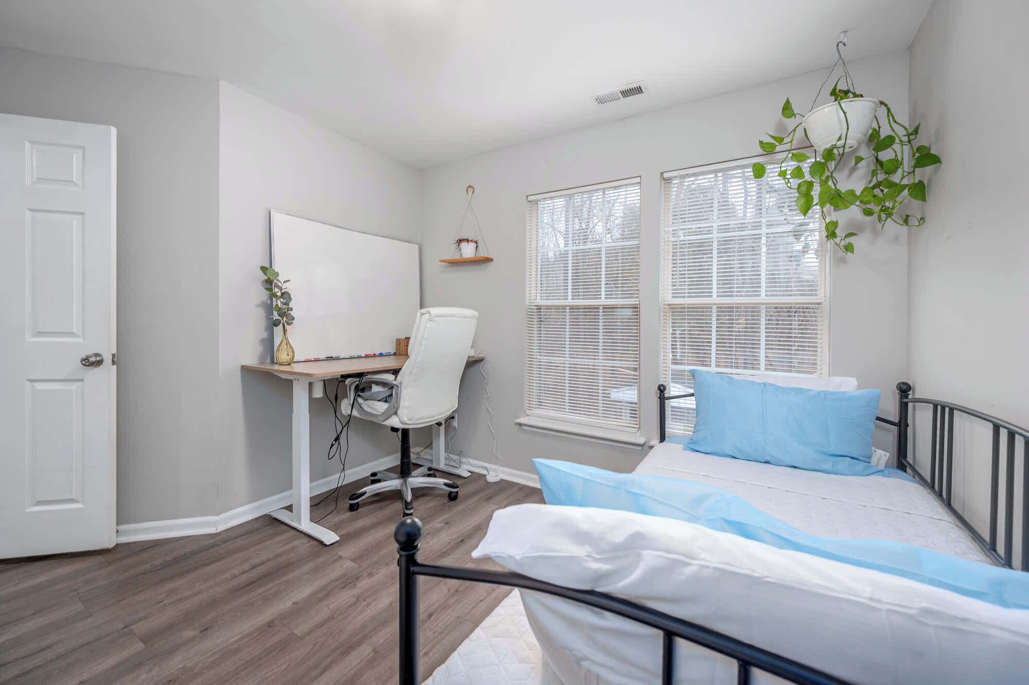 A bedroom with a bed, a desk, a chair, a whiteboard, and a window with blinds.