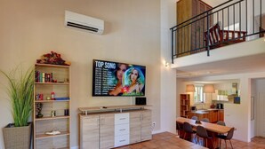 Un amplio salón con una mesa de comedor, un televisor mostrando una lista de canciones, una estantería y una planta grande en maceta.