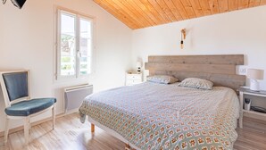 Un dormitorio con techo de madera, una cama con ropa de cama estampada, una silla, una mesita de noche con lámpara y una ventana con vistas.