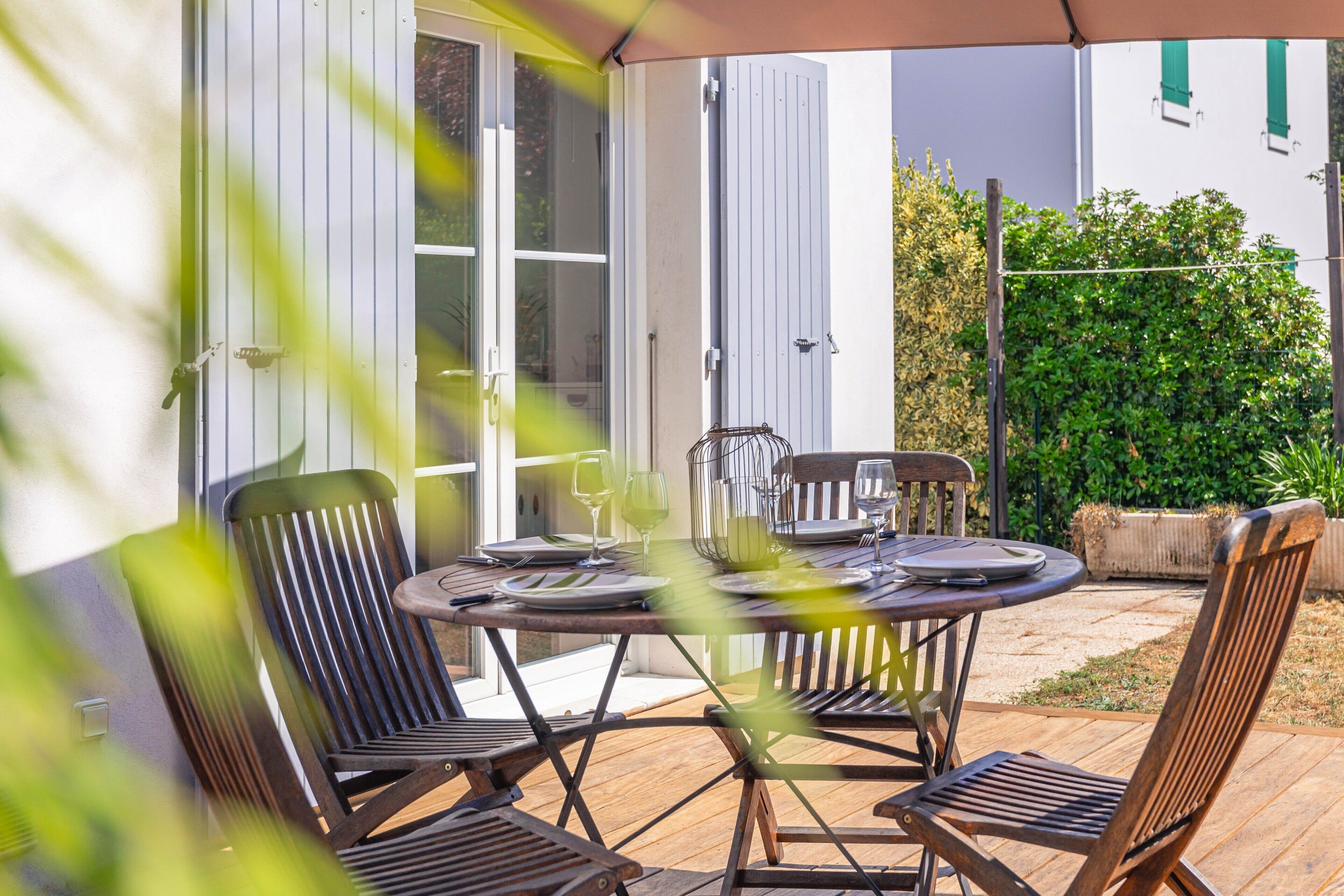 An outdoor patio with a round table and chairs, a small garden, and a white building with shutters.