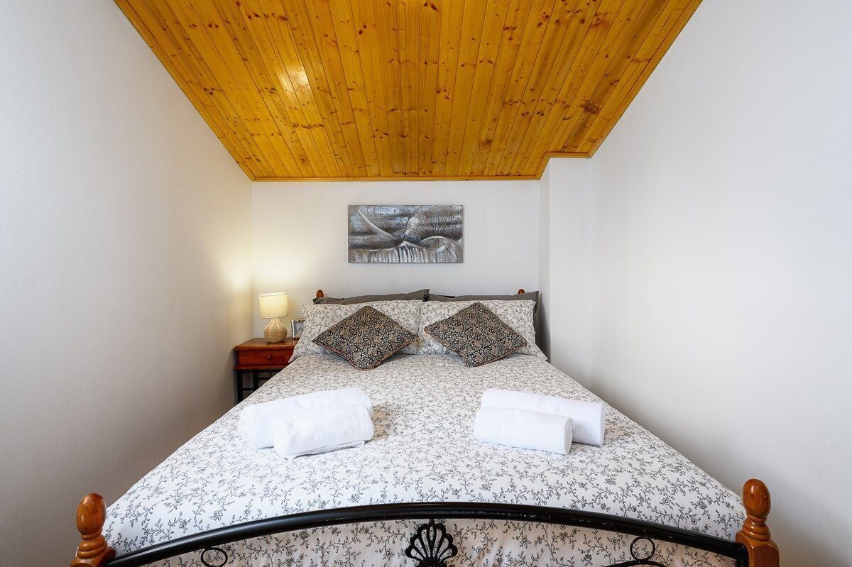 A neatly made bed with a floral bedspread, two decorative pillows, and two rolled towels on a wooden bed frame.