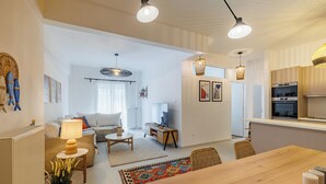 A modern living room with a sofa, coffee table, and lamp. There is a dining area with a wooden table and chairs. The room has a large window and a kitchen area with a refrigerator and oven. - Cozy and Spacious Apartment close to the Acropolis (Athina)