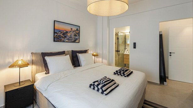 A hotel room with a large bed, bedside lamps, a painting on the wall, and a bathroom visible through an open door. - Cozy and Spacious Apartment close to the Acropolis (Athina)
