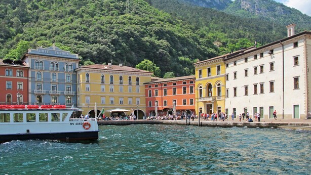 A waterfront hotel with colorful buildings, a boat docked nearby, and a mountainous backdrop. - Villa im Grünen in Riva del Garda mit Terrasse by Interhome (Riva del Garda)