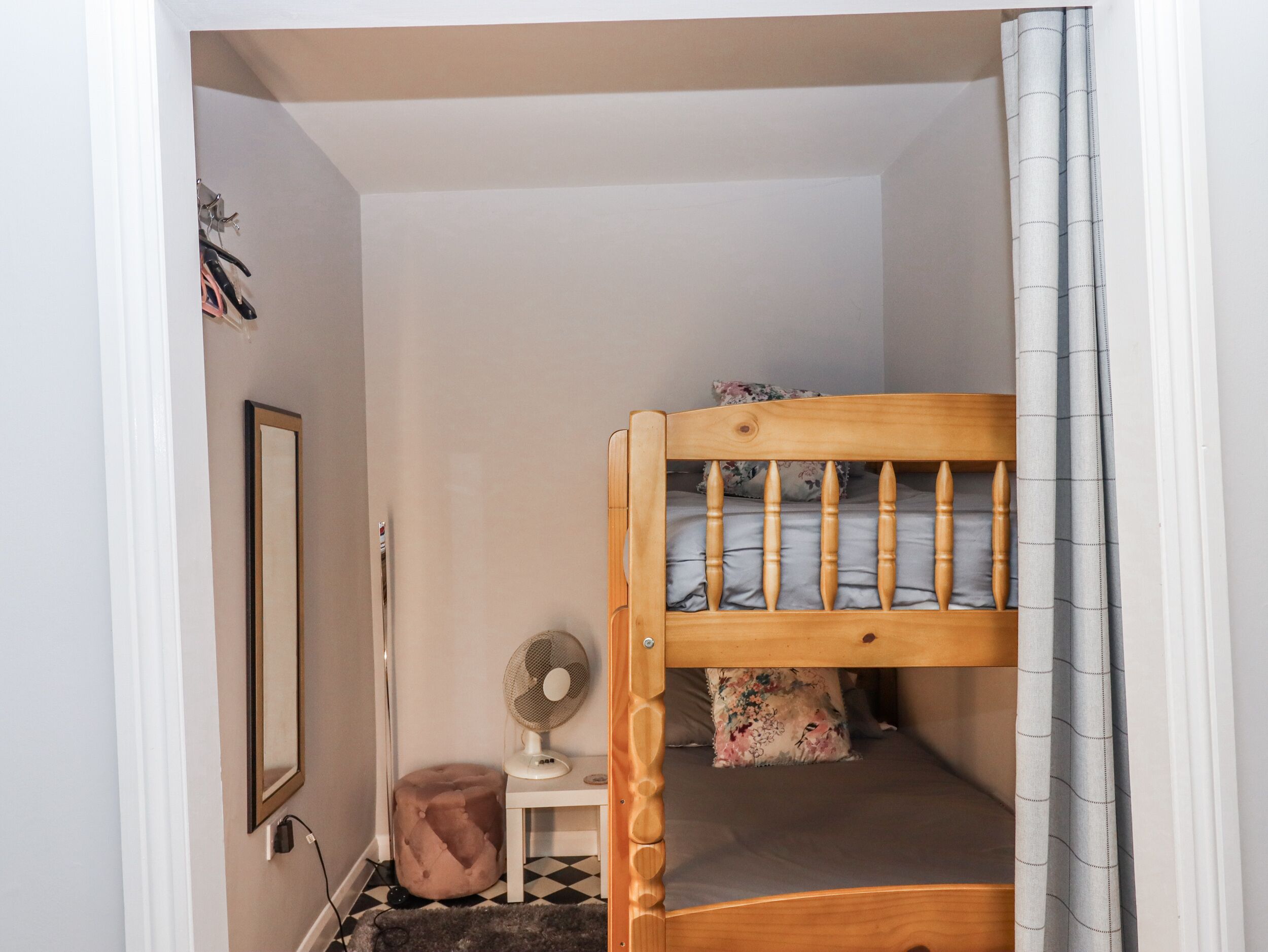Un dormitorio pequeño y ordenado con una cama litera, una mesita de noche con un ventilador, un taburete pequeño y un espejo en la pared.