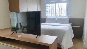 Habitación de hotel con cama, televisor sobre un mueble de madera, mesita de noche pequeña y ventana con vista a la ciudad.