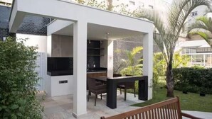 Un área de cocina al aire libre moderna con una mesa de comedor y sillas, rodeada de follaje y una estructura de edificio blanca.