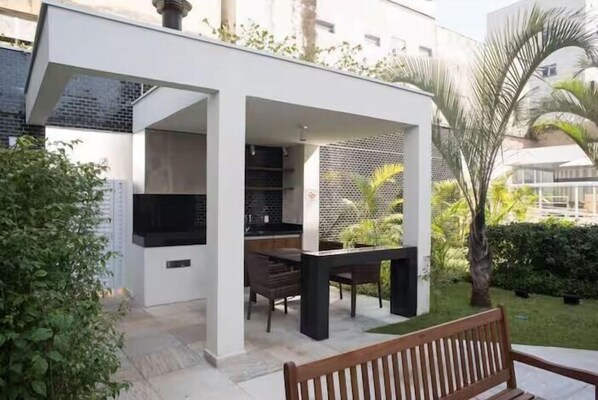 A modern outdoor kitchen area with a dining table and chairs, surrounded by greenery and a white building structure. - Conteporaneo 81 (São Paulo)