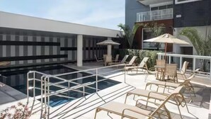 Área de piscina en la azotea con sillones de descanso y sombrillas, rodeada por un edificio moderno con balcones.