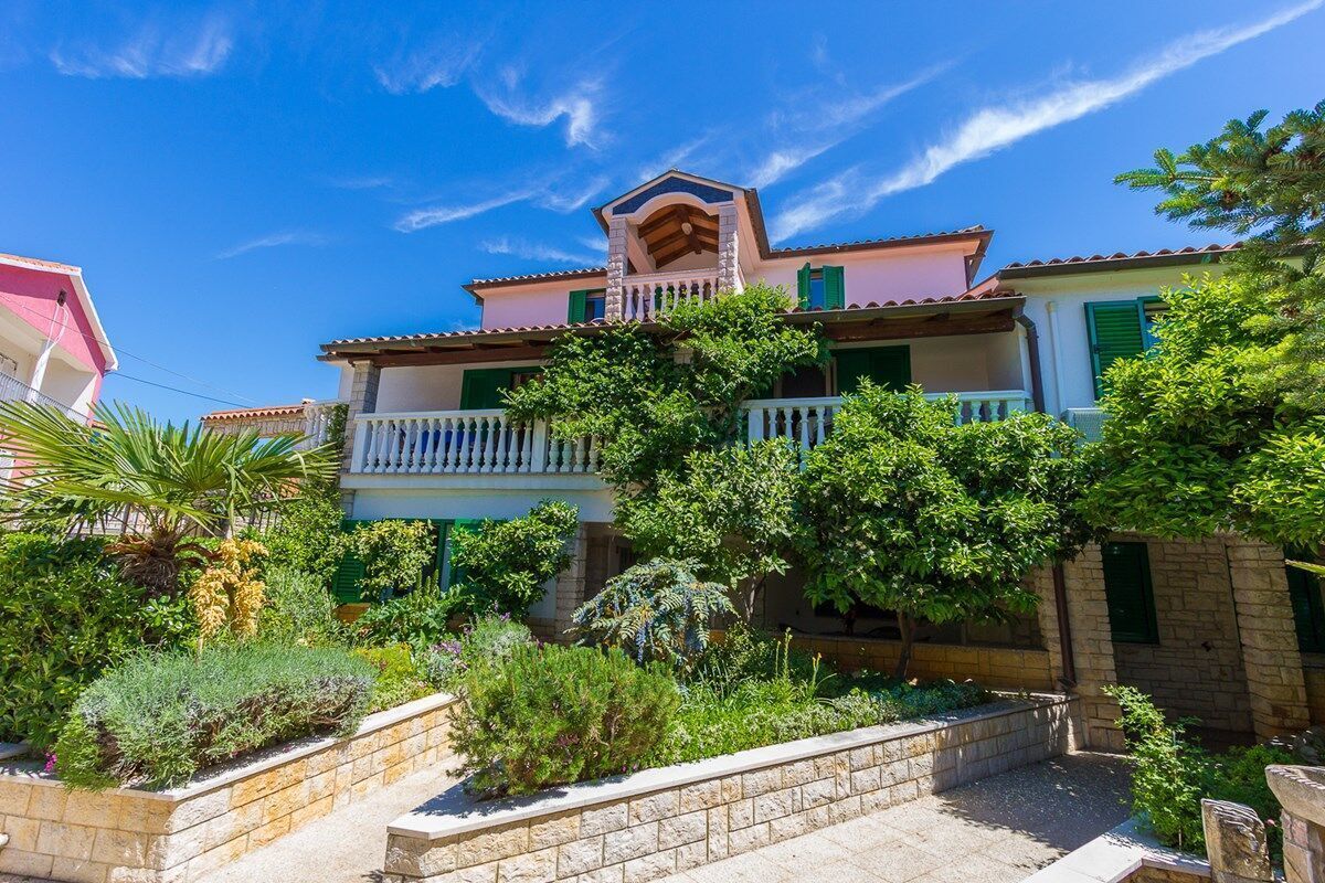 A two-story house with a balcony, surrounded by lush greenery and a stone wall.