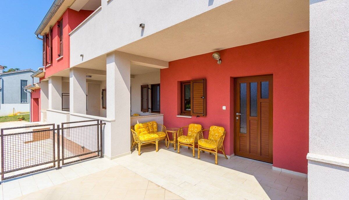 Un immeuble de deux étages avec une façade rouge et blanche, un balcon agrémenté de meubles en osier jaune et une entrée fermée par un portail.