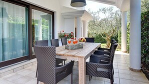 Een terras met een houten tafel en rieten stoelen, een hanglamp en een glazen deur die uitkomt in een tuin.