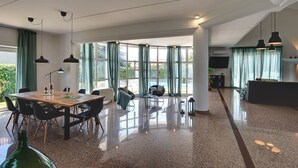 Een ruime woonkamer met een houten eettafel, zwarte stoelen, een bank en een televisie. Grote ramen met gordijnen zorgen voor natuurlijk licht en uitzicht op de omgeving.