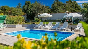 Una piscina con lettini bianchi e ombrelloni, immersa nel verde e circondata da un muro piastrellato.