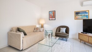 Ein Wohnzimmer mit beiger Couch, einem Glastisch, einem braunen Sessel, einem Fernseher auf einem Holzschrank und zwei gerahmten Bildern an der Wand.