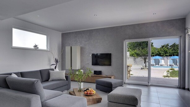 En moderne stue med grå sofaer, et sofabord med frugt, et fladskærms-tv og en skydedør af glas, der fører til et udendørs område med parasoller.