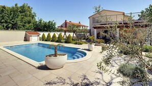 En swimmingpool med tilhørende terrasse, en stor hvid plantekasse og en bygning med to etager og balkon.
