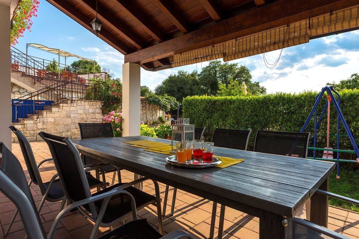 An outdoor patio with a wooden table and chairs, a metal railing, and a view of a garden and stairs.