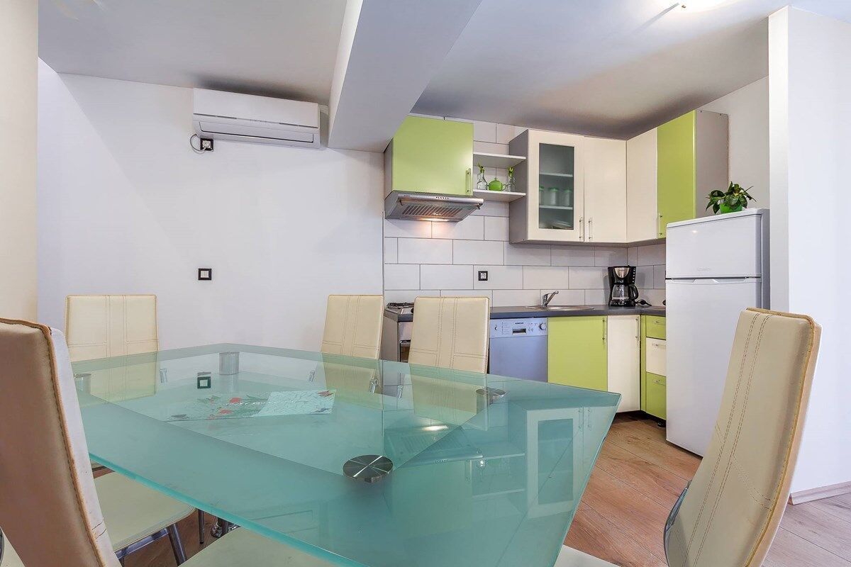 A modern kitchen with a glass dining table, beige chairs, and a refrigerator.