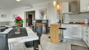 A modern kitchen with white cabinets, a gas stove, and a built-in oven. There is a dining area with a white table and chairs, and a vase with flowers on the table. - Villa Amber (V3551-K1) - Vodnjan (Vodnjan)