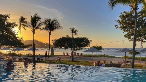 A pool area with lounge chairs and umbrellas, overlooking a beach with palm trees and a sunset. - Wake Up in Paradise, Amazing 3BDR Condo Near Beach (Coco)
