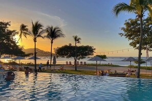 A pool area with lounge chairs and umbrellas, overlooking a beach with palm trees and a sunset. - Wake Up in Paradise, Amazing 3BDR Condo Near Beach (Coco)