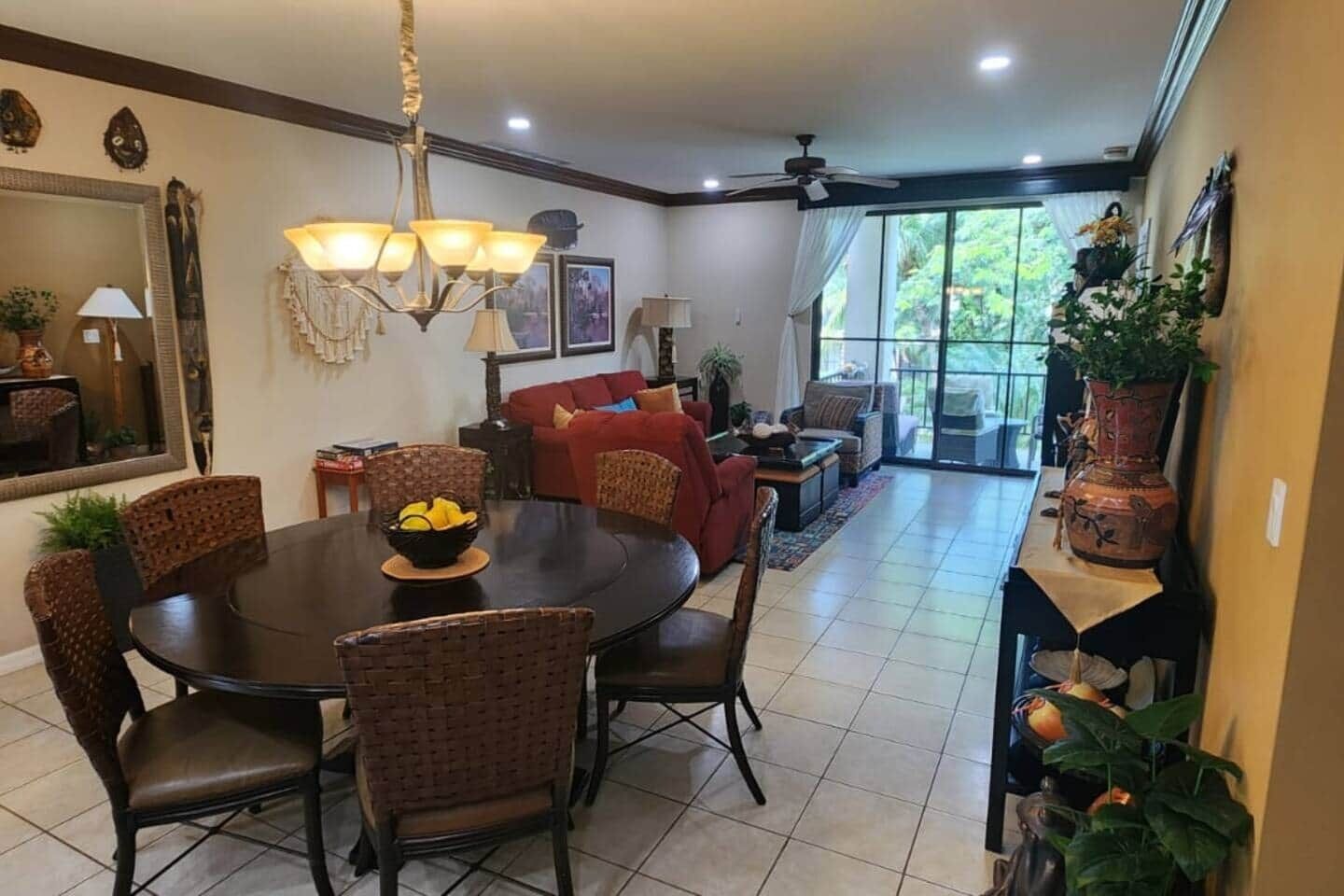 A dining area with a round table, wicker chairs, and a chandelier. There is a red sofa and a balcony with plants.