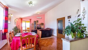 Ein Essbereich mit rosa Tischdecke, Holzkästen und einem schwarzen Sofa. Auf einer Arbeitsplatte steht ein Blumentopf und eine Tür mit Glasfenster.