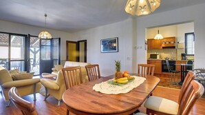 Un comedor con una mesa de madera, sillas y un centro de mesa con frutas. Se observa una cocina al fondo con armarios de madera y una lámpara colgante.