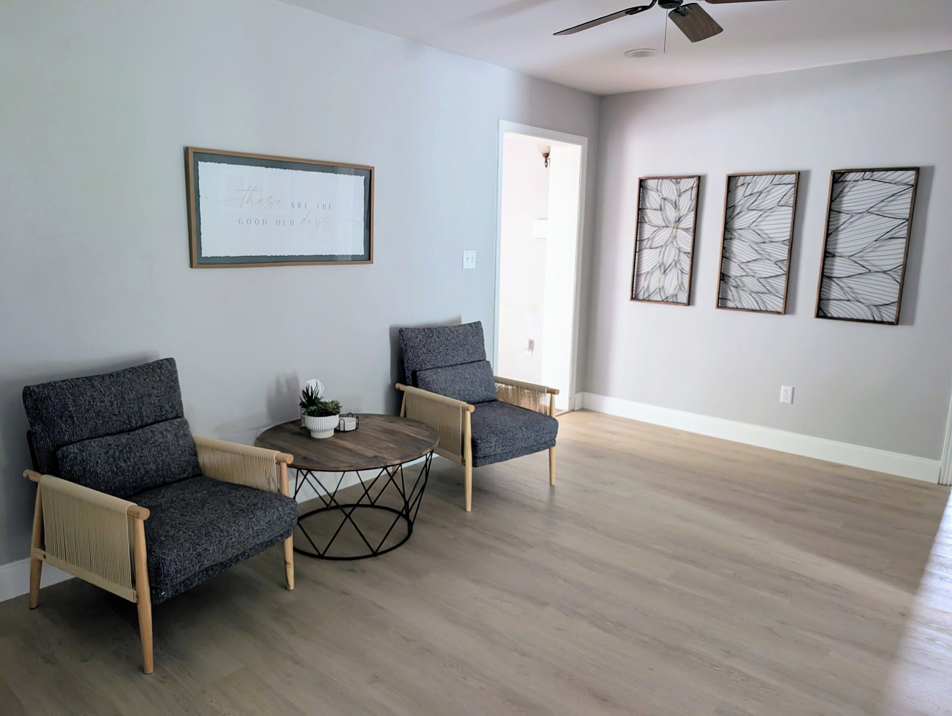 A modern living room with two grey armchairs, a round wooden coffee table with a plant, and three abstract wall art pieces.