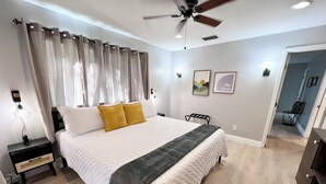 Un dormitorio con cama, mesita de noche, ventilador de techo y una puerta que da a otra habitación.
