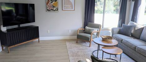 A modern living room with a flat-screen TV, a grey sofa, a wooden coffee table, and a grey armchair.