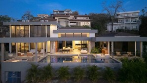Ein modernes, mehrgeschossiges Haus mit einem großen, verglasten Pool, Sitzgelegenheiten im Freien und einem gut beleuchteten Innenraum.