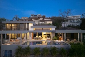 Ein modernes, mehrgeschossiges Haus mit einem großen, verglasten Pool, Sitzgelegenheiten im Freien und einem gut beleuchteten Innenraum.