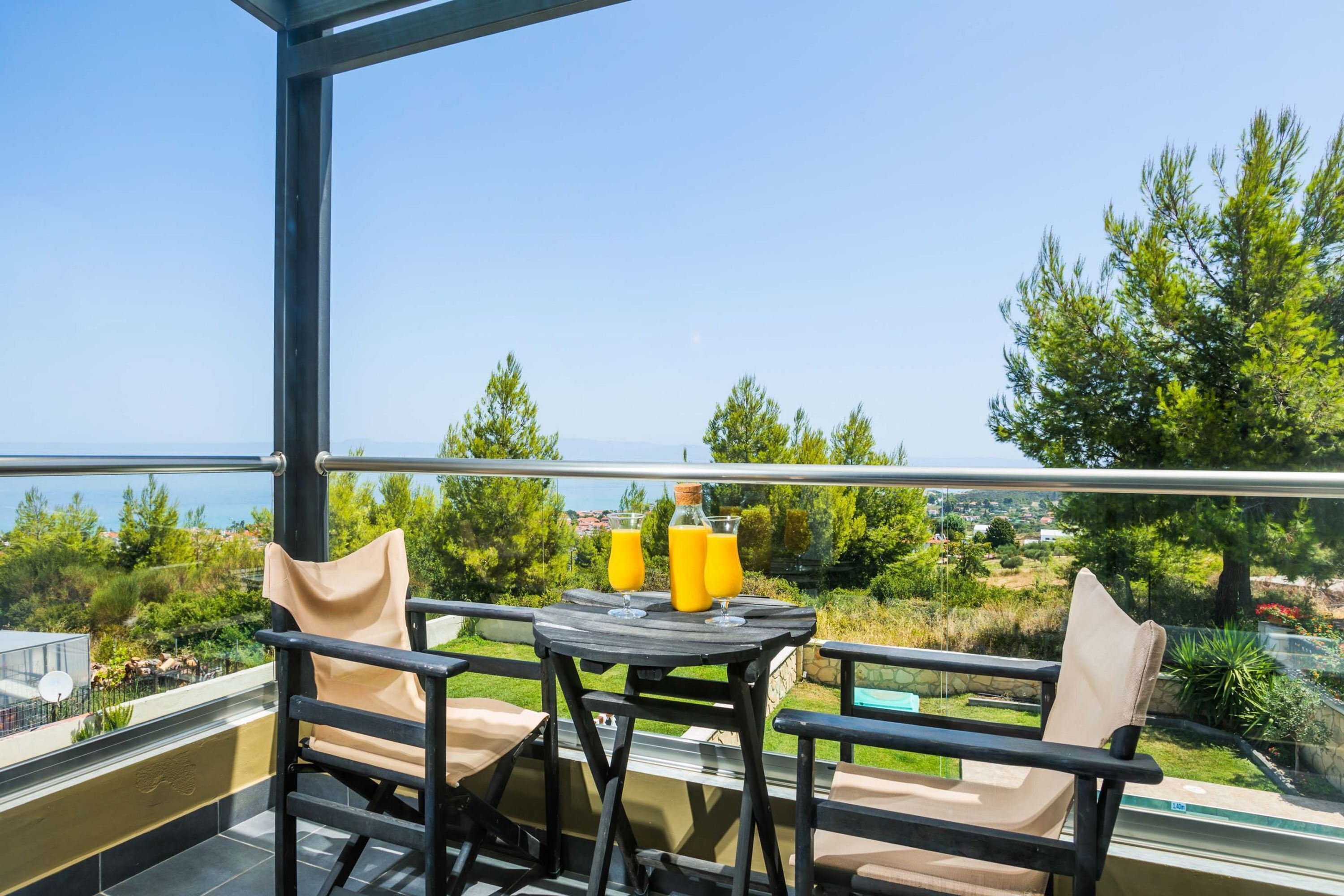 A balcony with two chairs and a table, two glasses of orange juice, and a view of the sea and greenery.