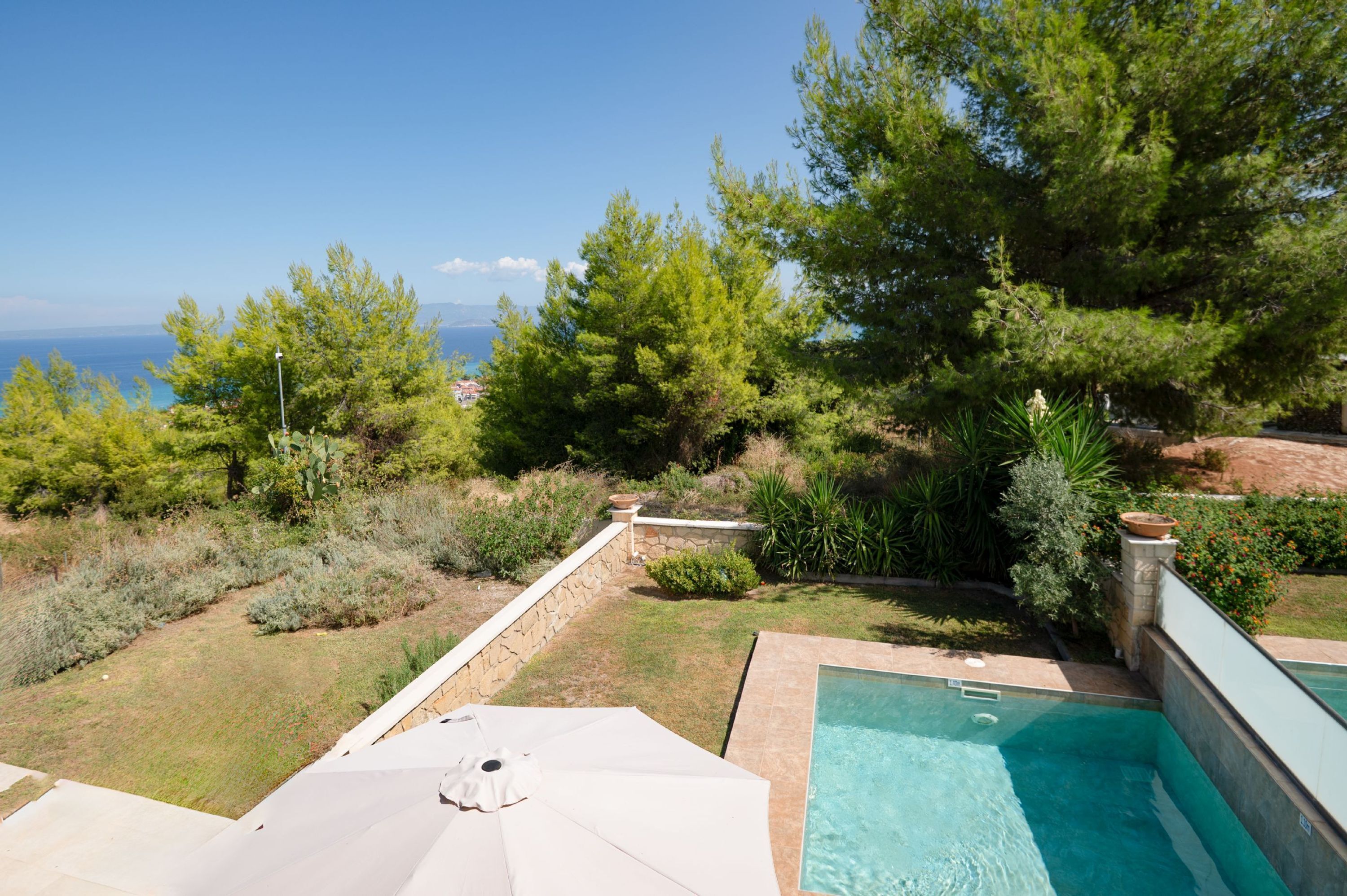 A pool area with a view of the sea, surrounded by greenery and a stone wall.