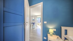 A room with a blue wall, a white bedside table with a lamp, a bed, and a door leading to a hallway. - CASA PIOPPI - Comfort and a beautiful view (Rapallo)