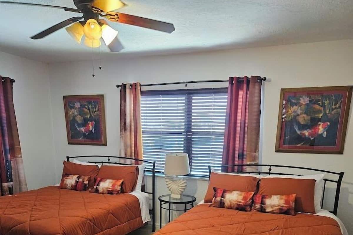 Een hotelkamer met twee bedden, een plafondventilator, een raam met zonwering en twee schilderijen aan de muur.