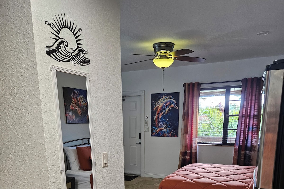 Een slaapkamer met een bed, een plafondventilator, een raam met gordijnen, een deur en een muur met een muurschildering van een zon en een golf.