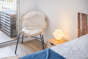 Ein Zimmer mit einem Bett, einem Sessel mit weichem Kissen, einem kleinen Nachttisch mit Lampe und Blick ins Freie durch eine Glastür.