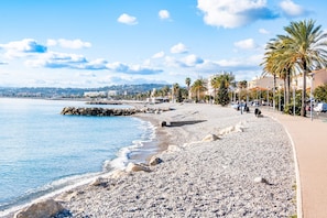A pebbled beach with a promenade, palm trees, and buildings in the background. - 'Couronne' Cros de Cagnes, Near Beaches, 2-Bedroom with Sea View, AC, and Parking (Cagnes-sur-Mer)