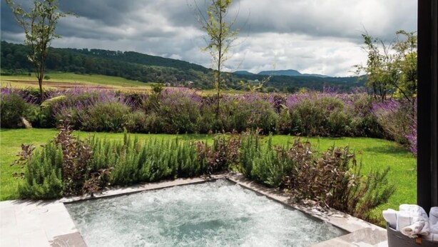 Area benessere con vasca idromassaggio, circondata da un giardino fiorito di fiori viola e vista su un paesaggio collinare.
