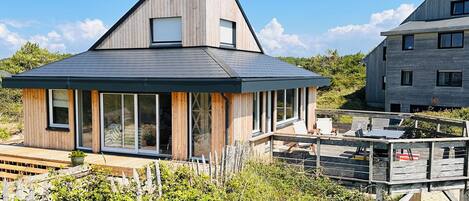 A modern house with a sloped roof, large windows, and a deck with outdoor furniture.