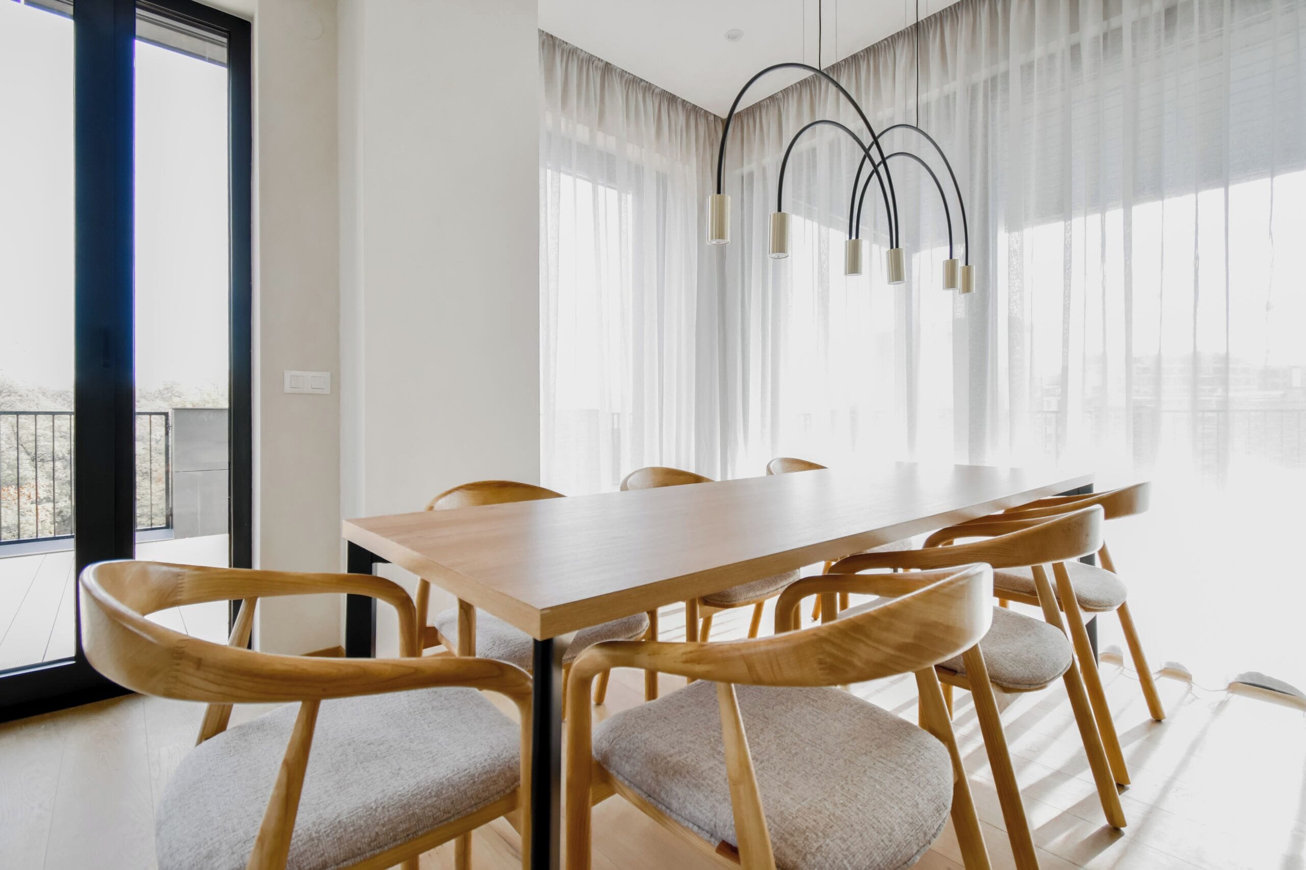 A modern dining area with a wooden table and chairs, large windows with sheer curtains, and a unique ceiling light fixture.