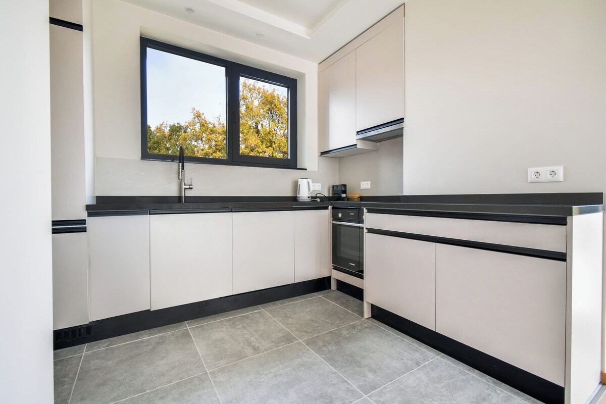 A modern kitchen with white cabinets, a black countertop, a window with a view of trees, and a built-in oven.