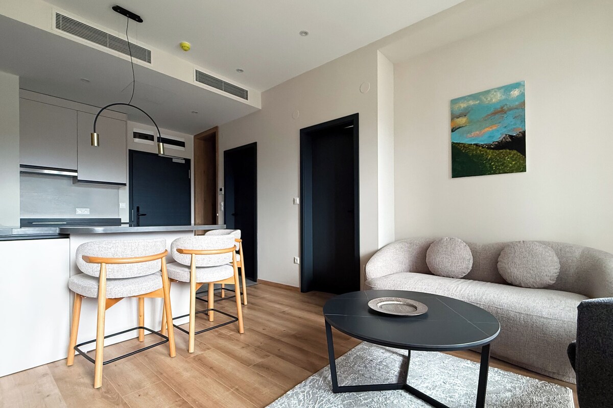 A modern living space with a kitchen, a dining area with two bar stools, a round coffee table, a sofa with round cushions, and a painting on the wall.